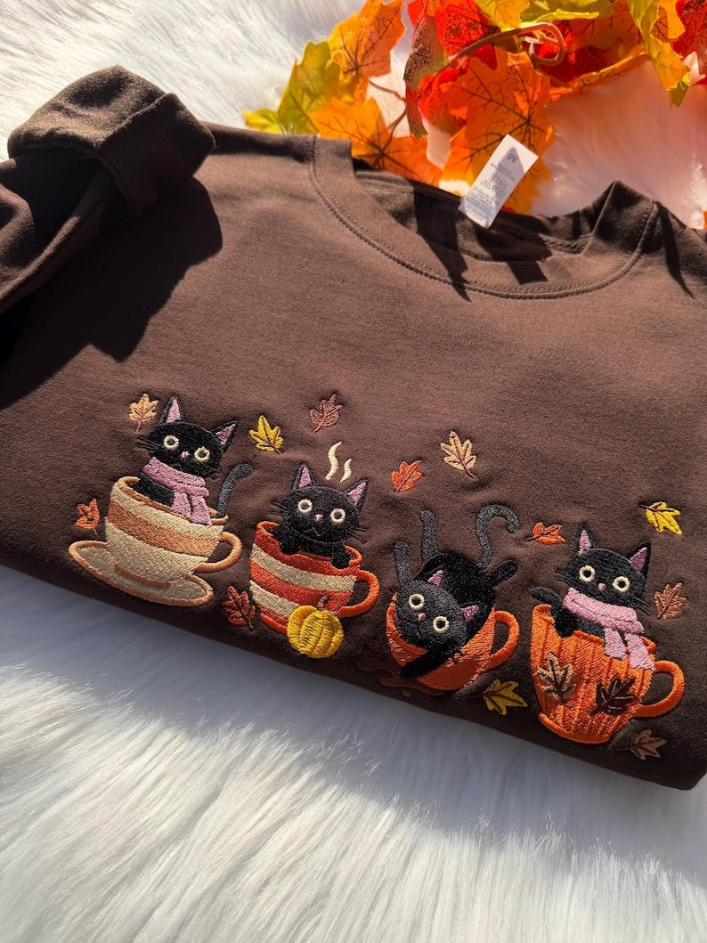 Detailed view of embroidered black cats in mugs with pumpkins on brown sweatshirt