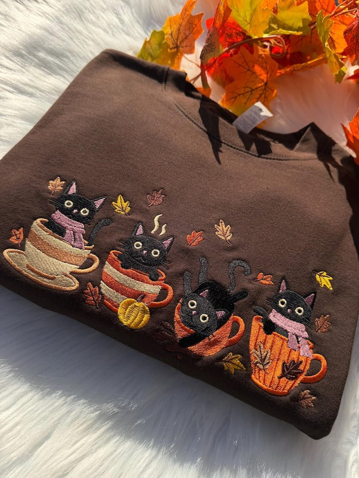 Folded brown sweatshirt with embroidered black cats in mugs and fall leaves