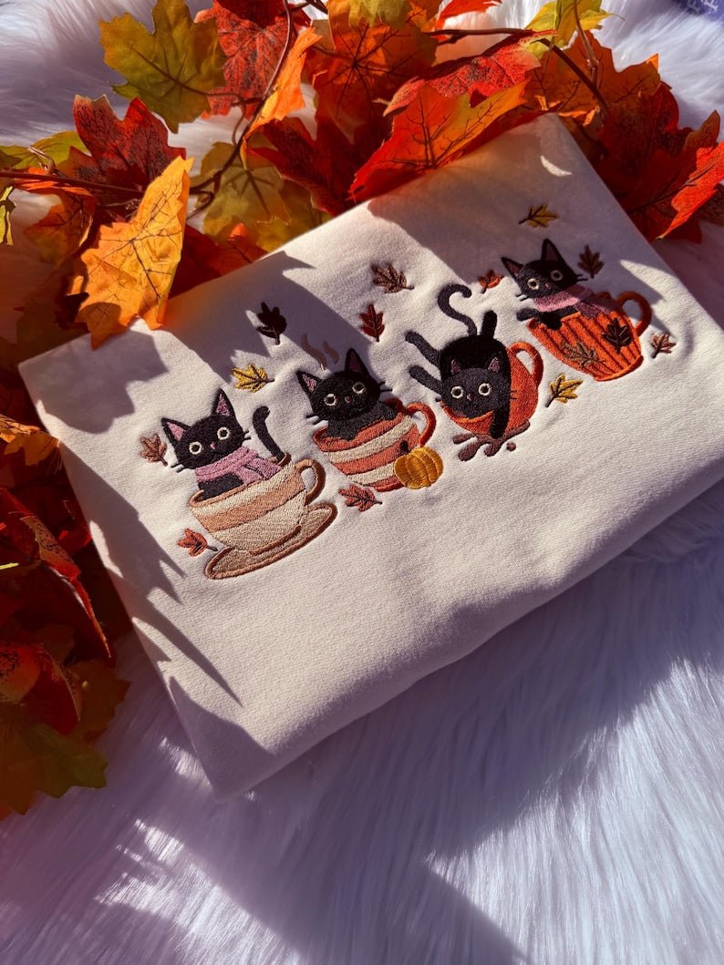 Folded milkshake sweatshirt with embroidered black cats in mugs, styled with autumn leaves