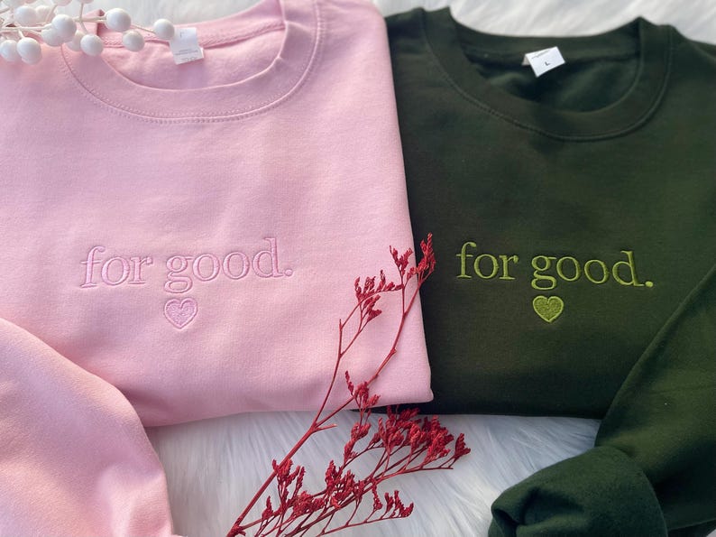 Flatlay of pink and green sweatshirts with Wicked-inspired “For Good.” embroidery