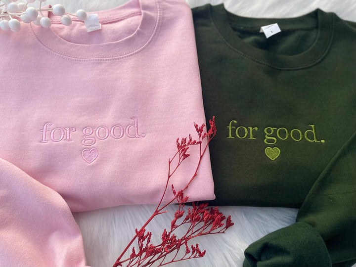 Flatlay of pink and green sweatshirts with Wicked-inspired “For Good.” embroidery